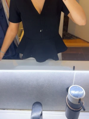 Svart peplumtop med v-ringning - Snygg svart peplumtop med v-ringning och korta ärmar. Toppen har en markerad midja och volang nedtill som ger en feminin siluett. Perfekt att styla med både jeans och kjol för en trendig look.