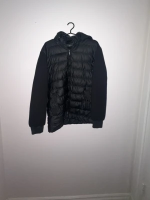 Svart dunjacka med huva - Snygg svart dunjacka med quiltat mönster och huva. Jackan har svarta ärmar i mjukt material och två fickor framtill med knappdetaljer. Perfekt för kalla vinterdagar och har en modern, ungdomlig look. Lite små defekter såsom att förgen har lossnat på lite olika ställen efter ganska mycket användning 