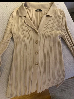 Beige ribbad kofta från Gina Tricot - Snygg beige ribbad kofta från Gina Tricot med knappar framtill och krage. Långärmad modell i mjukt, stretchigt material som sitter skönt på kroppen. 