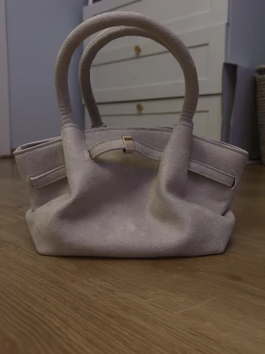 Beige mocka handväska med gulddetalj - Stilren beige handväska i mjuk mocka med två rejäla handtag och en snygg guldfärgad spänndetalj framtill. Väskan har en klassisk, rymlig form och passar perfekt för dig som gillar en enkel men trendig look.