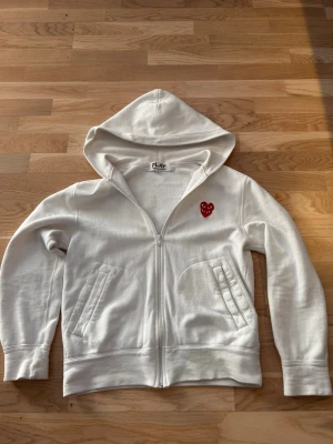 Vit hoodie från Comme des Garçons Play - Vit hoodie med dragkedja från Comme des Garçons Play. Klassisk röd hjärtlogga med ögon på bröstet, två snedställda fickor fram och huva. Tillverkad i mjuk bomull som känns skön mot huden. Perfekt för en clean och stilren look.
