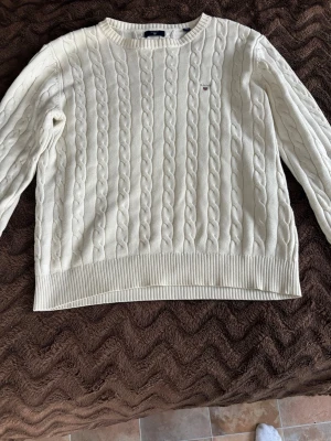 Vit kabelstickad tröja från Gant - Kabelstickad vit tröja från Gant med rund halsringning och diskret broderad logga på bröstet. Tröjan har ribbade muddar vid ärmslut och nederkant, och är tillverkad i mjukt material som ger en klassisk och stilren look.