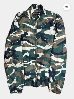Camo vindjacka från Woolrich - Snygg vindjacka från Woolrich i klassiskt camomönster med gröna, beige, vita och mörkblå toner. Jackan har dragkedja framtill, elastiska muddar och lätt material som passar perfekt för blåsiga dagar. Märkeslogga på bröstet och ärmen ger en extra touch. Det är i bra skick 9/10, priset kan diskuteras vid snabba affär 