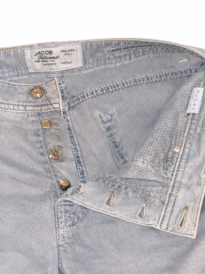 Jacob cohen Jeans - Säljer ett par ljusblå jeans från Jacob med tapered passform och klassisk femficksdesign. Jeansen har knäppning med metallknappar och subtila slitningar för en avslappnad look. Tillverkade i Italien och har snygga detaljer vid fickorna.