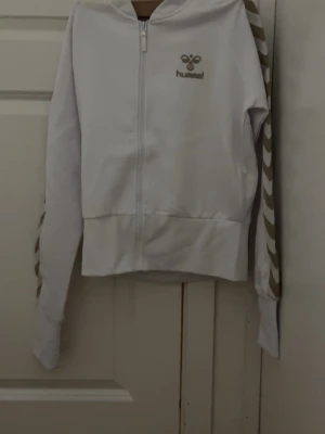 Vit hoodie med dragkedja från Hummel - Vit hoodie från Hummel med dragkedja framtill och beigea chevronränder längs ärmarna. Hummel-logga på bröstet. Klassisk huva och ribbade muddar. Perfekt för en sportig och avslappnad stil.