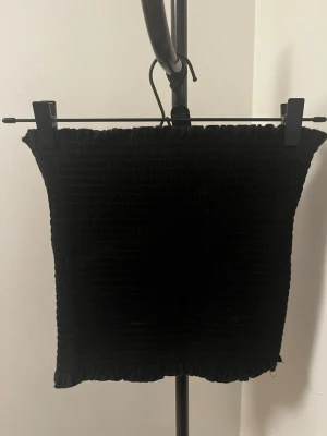 Svart bandeau tubtopp från H&M - Svart bandeau tubtopp från H&M med smockad struktur och volangkant upptill och nedtill. Toppen är ärmlös och har en tight passform, perfekt att styla till jeans eller kjol.