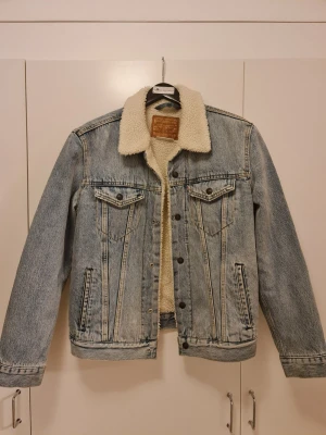 Levi's fodrad jeansjacka blå - Klassisk blå jeansjacka från Levi's med mysigt vitt teddyfoder och krage. Jackan har två bröstfickor med lock, sidofickor och knäppning framtill. Perfekt för dig som vill ha en cool och tidlös look. Jackan är i mycket bra skick så passar på att fynda denna tidlösa jacka!