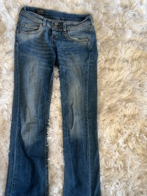 Blå bootcut jeans från Lee - Säljer ett par klassiska blå bootcut jeans från Lee med snygg slitning framtill och bakfickor. Jeansen har låg midja, fem fickor och är gjorda i mjuk denim med lite stretch. Perfekta för en avslappnad och trendig look.
