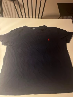 Mörkblå t-shirt från Ralph Lauren - Klassisk mörkblå t-shirt från Ralph Lauren med röd broderad logga på bröstet. T-shirten har rund halsringning och korta ärmar, tillverkad i mjuk bomull för en skön känsla. Perfekt för en avslappnad och stilren look.