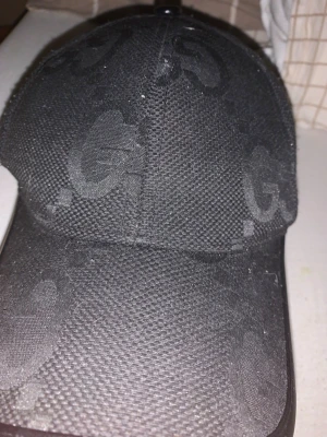 Svart keps från Gucci med GG-mönster - Snygg svart keps från Gucci med diskret GG-monogram över hela kepsen. Justerbar rem baktill för perfekt passform. Tillverkad i textilmaterial med klassisk böjd skärm. Perfekt accessoar för en trendig streetstyle-look.