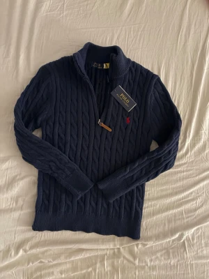 Mörkblå kabelstickad tröja Polo Ralph Lauren - Mörkblå kabelstickad tröja från Polo Ralph Lauren med halv dragkedja och klassisk röd logga broderad på bröstet. Tröjan har hög krage och ribbade muddar. Perfekt för dig som gillar stilren och tidlös design.
