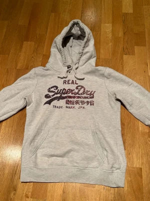 Grå hoodie från Superdry - Snygg ljusgrå hoodie från Superdry med stor logga och japansk text framtill. Klassisk känguruficka, dragsko i huvan och ribbade muddar. Perfekt för en avslappnad och trendig stil. Mjuk bomullsblandning som känns skön mot huden.