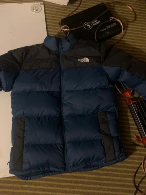 Blå dunjacka från The North Face - Säljer en blå dunjacka från The North Face med svarta detaljer på axlar och fickor. Jackan har hög krage, dragkedja framtill och två stora fickor. Perfekt för kalla dagar och riktigt snygg streetstil.