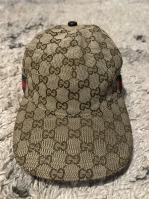 Gucci beige GG-mönstrad keps -  Säljer en beige keps från Gucci med det klassiska GG-monogrammönstret över hela kepsen. Kepsen har en böjd skärm och justerbar rem bak. På sidorna finns de ikoniska gröna och röda ränderna. Materialet är canvas med detaljer i bomull.