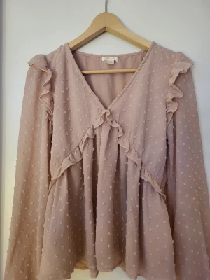 Rosa volangblus med prickar - Söt ljusrosa blus med volangdetaljer och små prickar i tyget. Blusen har v-ringning, transparenta långärmar och ett luftigt, flowy fall. Perfekt för dig som gillar romantisk och feminin stil. Materialet känns lätt och skönt mot huden.