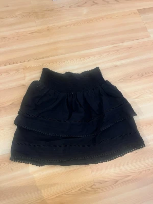 Svart volangkjol - Kort svart kjol med bred resår i midjan och flera lager volanger. Kjolen har dekorativa spetsdetaljer längs volangkanterna och är perfekt för en trendig och avslappnad look.