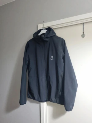 Svart vindjacka från Haglöfs - Snygg svart vindjacka från Haglöfs med huva och dragkedja framtill. Jackan har två sidofickor med dragkedja och diskret logga på bröstet. Perfekt för blåsiga dagar och enkel att matcha med olika outfits.