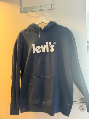 Mörkblå hoodie från Levi's - Snygg mörkblå hoodie från Levi's med stor vit logga på bröstet. Klassisk modell med huva och känguruficka framtill. Mjuk bomullsblandning som är skön att ha på sig. Perfekt för en avslappnad stil.