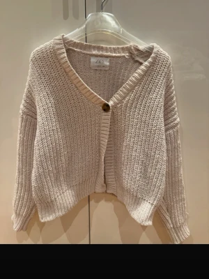 Beige stickad kofta från Zara - Mysig beige stickad kofta från Zara med grovstickad struktur och en knapp framtill. Koftan har en avslappnad, lite kortare passform och långa ärmar med ribbade muddar. Perfekt att slänga över en topp för en chill look.