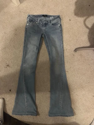 Blå bootcut jeans från true religion - Snygga blå bootcut jeans från true religion med klassisk femficksdesign och låg midja. Jeansen har en lätt tvättad look och utsvängda ben, perfekta för en avslappnad och trendig stil. Materialet är bomullsdenim med lite stretch för extra komfort.