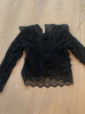 Svart spetsblus med lång ärm - Snygg svart spetsblus med långärmad design och detaljerad spets över hela plagget. Blusen har rund halsringning och dekorativa kanter nertill samt vid ärmslut. Perfekt för dig som gillar en elegant och trendig stil.