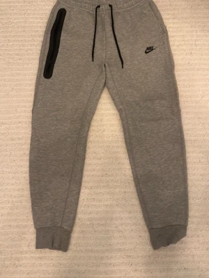 Nike tech byxor gråa knappt använda som helt ny  - Snygga gråa mjukisbyxor från Nike med svart logga på vänster ben och svarta dragsnören i midjan. Ena fickan har en cool svart dragkedja. Byxorna har muddar vid bensluten och är gjorda i mjukt sweatshirtmaterial. Perfekta för chill eller träning.