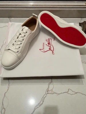 Vita Christian Louboutin sneakers - Snygga vita sneakers från Christian Louboutin med klassisk röd sula och stilren design. Skorna är låga och har vita skosnören samt ovandel i slätt läder. Perfekta för dig som vill ha en exklusiv och trendig look.