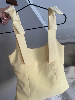 Gul linnetopp med rosetter - Supersöt ljusgul linnetopp med breda axelband som knyts i stora rosetter. Toppen har en rak, croppad passform och är perfekt för varma dagar. Materialet känns mjukt och lätt, och färgen ger en fräsch vibe till din outfit.