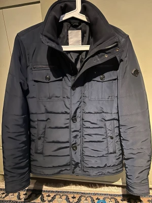 J.Lindeberg fieldjacka S (Bailey Poly) - Snygg mörkblå fieldjacka från J. Lindeberg med quiltad design, hög krage och fyra fickor framtill. Jackan har en diskret logotyp på ärmen och stängs med både dragkedja och knappar. Perfekt för vårdagae och ger en stilren look.