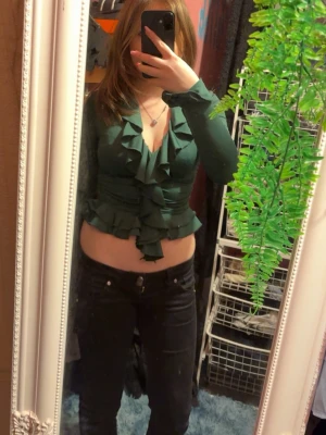 Mörkgrön volangblus med v-ringning - Trendig mörkgrön blus med djup v-ringning och volangdetaljer längs halsringningen och framtill. Figurnära passform och bred resår i midjan som framhäver siluetten. Långärmad modell i mjukt, stretchigt material. Fin för fester också💕