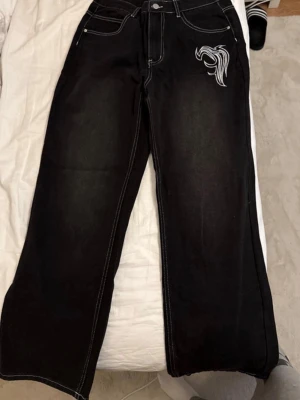 Svarta vida jeans med vit brodyr - Säljer ett par svarta jeans med bred passform och vit kontrastsöm. Jeansen har en unik vit brodyr på ena fickan framtill och klassiska bakfickor. Materialet är denim och modellen är rak och avslappnad, perfekt för en trendig streetwear-look.