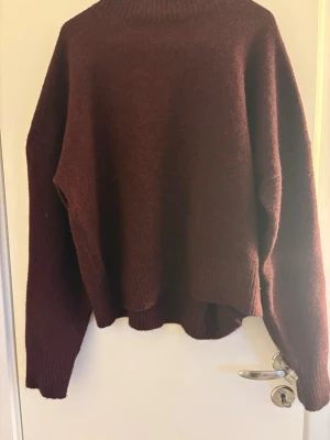 Vinröd stickad tröja Samsa Collection - Mysig vinröd stickad tröja från Samsa Collection med hög hals och avslappnad passform. Tröjan har långa ärmar och ribbade muddar vid hals, ärmslut och nederkant. Perfekt för kalla dagar och enkel att styla till olika outfits.