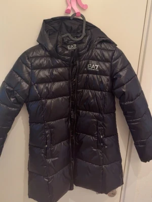 Svart dunjacka EA7 Emporio Armani - Snygg svart dunjacka från EA7 Emporio Armani med huva och dragkedja. Jackan har quiltad design och logga på bröstet. Perfekt för kalla dagar och ger en sportig look.