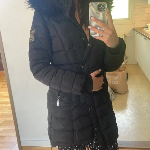 Svart dunjacka med fuskpäls från Chiara Forthi - Säljer en svart lång dunjacka från Chiara Forthi med fuskpäls på huvan och dragkedja framtill. Jackan har quiltad design, lång ärm och snygg badge på ärmen. Perfekt för kyliga dagar.