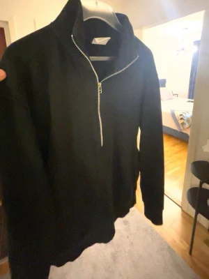Svart half-zip hoodie - En stilren svart hoodie med half-zip dragkedja framtill och hög krage. Hoodien har långa ärmar och ribbade muddar vid ärmslut och nederkant. Perfekt för en avslappnad look och enkel att matcha med andra plagg.