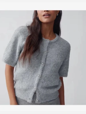 Grå kortärmad kofta - Mysig grå kofta med korta ärmar och rund hals. Koftan är gjord i ett fluffigt, stickat material som känns både trendigt och bekvämt. Perfekt att styla över ett linne eller t-shirt för en chill look.