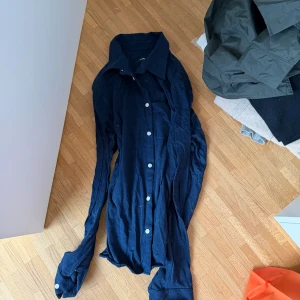 Mörkblå skjorta från J.Lindeberg - Snygg mörkblå skjorta från J.Lindeberg med klassisk krage och vita knappar. Skjortan är långärmad och tillverkad i mjuk bomull, perfekt för en stilren look. Passar dig som gillar tidlös design och enkelhet.