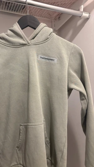 Ljusgrå hoodie från Peak Performance - Snygg ljusgrå hoodie från Peak Performance med broderad logga på bröstet. Klassisk modell med huva och känguruficka framtill. Tillverkad i mjuk bomullsblandning som känns skön mot huden. Perfekt för en avslappnad och stilren look.