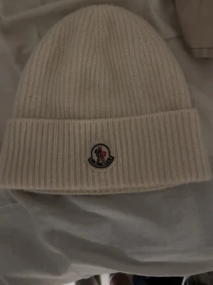 Vit ribbstickad mössa från Moncler - Säljer en stilren vit ribbstickad mössa från Moncler med uppvikt kant och klassisk Moncler-logga framtill. Mössan är gjord i mjukt material och har en clean look som passar perfekt till kalla dagar.