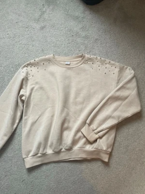 Beige sweatshirt med pärldetaljer - Trendig beige sweatshirt med rund hals och långa ärmar. Dekorerad med vita pärlor på axlarna för en extra snygg touch. Tillverkad i mjukt material och har ribbade muddar vid ärmslut och nederkant. Perfekt för dig som vill ha en enkel men ändå unik tröja. lite småfläckar på en ärmen som syns i bild. men syns ej tydligt