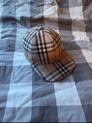 Burberry keps - Säljer en klassisk beige keps från Burberry med det ikoniska rutiga mönstret i svart, vitt och rött. Kepsen har böjd skärm och justerbar rem bak med Burberry London England-märke. Perfekt accessoar för en trendig och stilren look.