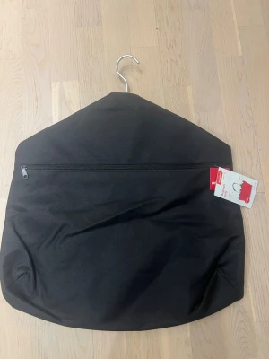 Svart kostympåse från Rusta - Säljer en svart kostympåse från Rusta med dragkedja framtill och praktisk hängare. Tillverkad i slitstarkt tyg och perfekt för att skydda kläder under transport eller förvaring. Enkel och stilren design med röd etikett på sidan.