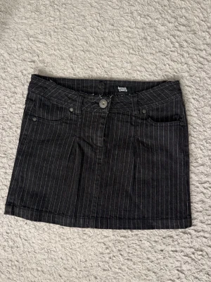 Svart kortkjol med vita ränder - Säljer denna snygga vintage kjol från med tunna vita ränder. Kjolen har klassisk jeanslook med fickor fram och bak, bälteshällor och knappstängning. Perfekt för dig som gillar en edgy och stilren vibe.