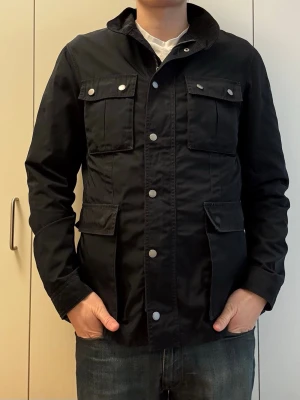 Svart utilityjacka med fickor - Svart utilityjacka med fyra stora fickor framtill och silverfärgade knappar. Jackan har hög krage och är tillverkad i ett slitstarkt material, perfekt för en stilren och funktionell look. Långärmad och med rak passform. Längd: 72cm, p2p: 50cm