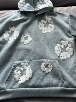  Denim tears Hoodie grå - Snygg ljusgrå hoodie med stora och små vita blommönster över hela tröjan, inklusive huvan och ärmarna. Klassisk känguruficka framtill och mjukt material som känns skönt mot huden. Perfekt för dig som gillar unika detaljer och streetwear-vibe. Passar även m. Säljer den för att jag har fått den och inte kommit till någon användning.