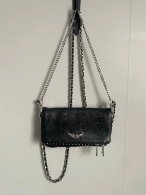 Zadig & Voltaire väska - Snygg och tidlös väska från Zadig & Voltaire i svart läder. Klassisk modell med silverfärgad kedjerem och märkets ikoniska vinglogga framtill. Kan bäras både som axelväska och crossbody och passar lika bra till vardag som fest. Väskan är i mycket fint skick, sparsamt använd och utan tydliga tecken på slitage. Lädret är fint och kedjan i bra skick. Original dustbag medföljer.