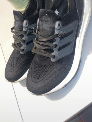 Svarta Adidas Ultraboost sneakers - Svarta Adidas Ultraboost sneakers med stickad ovandel och klassiska tre ränder i plast på sidorna. Skorna har svart snörning och en vit, tjock Boost-sula för extra komfort. Perfekta för dig som gillar sportig och stilren look.