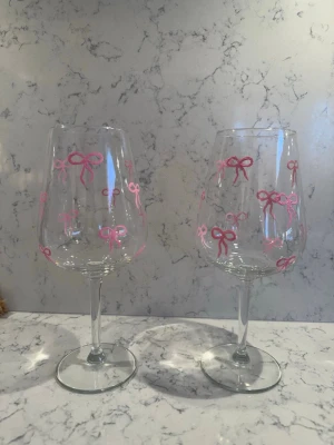 Vinglas med rosa rosetter - Två vinglas i klart glas dekorerade med handmålade rosa rosetter runt hela kupan. Glasen har klassisk form med lång smal fot och bred kupa, perfekta för att ge en söt och personlig touch till dukningen.
