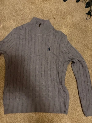 Ljusgrå kabelstickad tröja Polo Ralph Lauren - Säljer en ljusgrå kabelstickad tröja från Polo Ralph Lauren med halv dragkedja och hög krage. Tröjan har det klassiska broderade logotypen på bröstet och ribbade muddar. Perfekt för dig som gillar stilren och tidlös design.