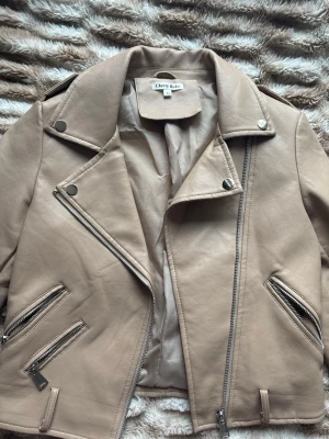 Beige skinnjacka från Cherry Koko - Snygg beige skinnjacka från Cherry Koko med klassisk bikerstil. Jackan har flera silverfärgade dragkedjor, breda slag och axelvaddar. Materialet är mjukt och jackan har en kort, figurnära passform. Perfekt för att lyfta din outfit med en cool vibe. Pris kan diskuteras